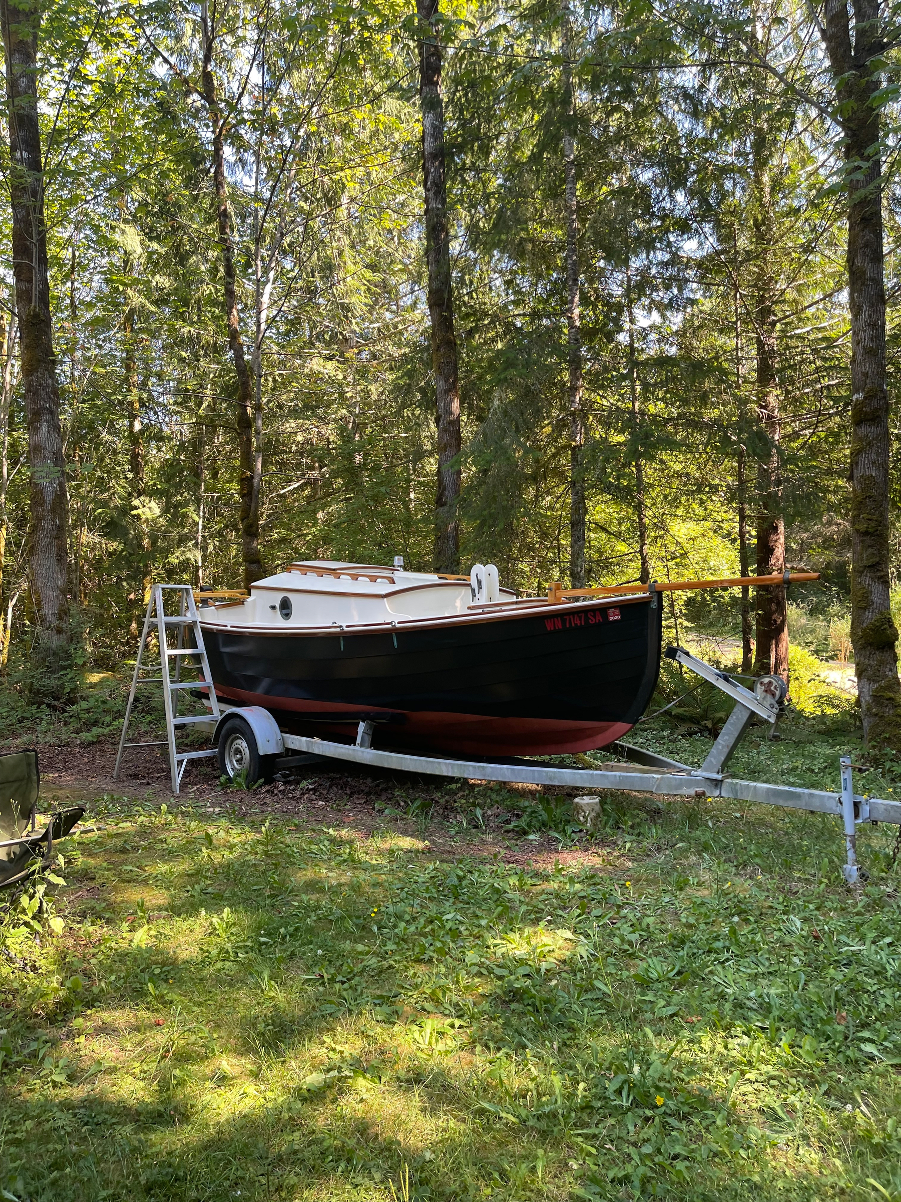 A small boat on a trailer is parked in a lush, wooded area with tall trees.